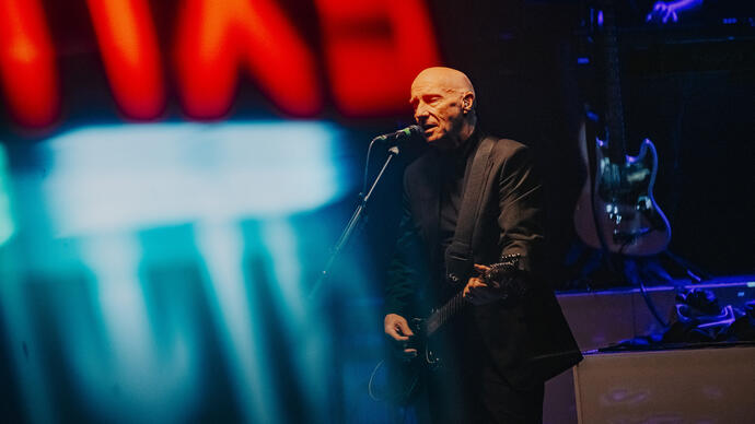 a photo of Midge Ure in a suit playing the guitar under the haze of a red and blue neon sign. 