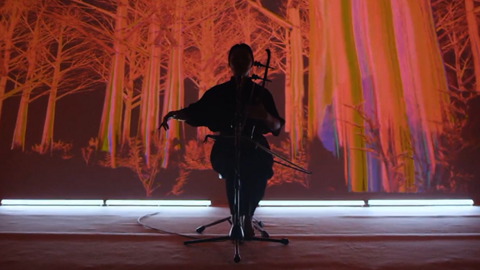 Silhouette of a person sitting and playing the cello infront of a screen with red trees on it