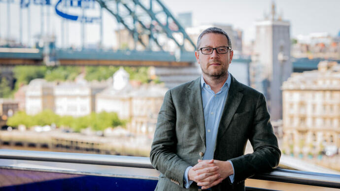 A man in a suit wearing glasses stands on a balcony with views of london behind him.