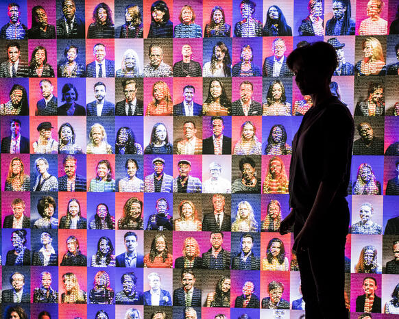 Digital installation of portraits of hundreds of people on a pink or purple ton background with the silhouette of a person standing in front of the installation on the right. 