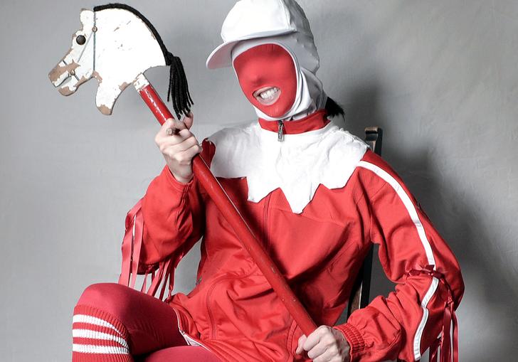 Gazelle Twin wearing a jester costume with a hobby horse, seated on a chair