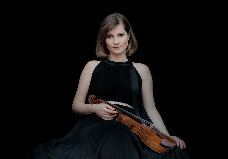 Lisa Batiashvili holding her violin and wearing a black dress standing in front of a black background.