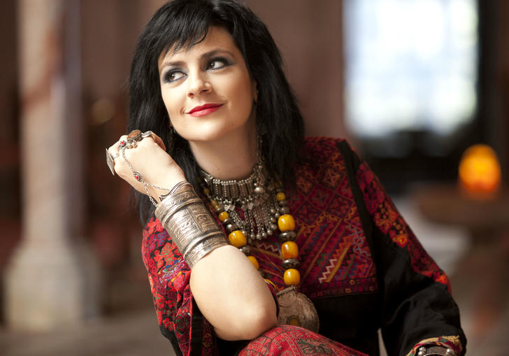 Rim Banna wearing a red dress and large jewellery. She is leaning on her elbow and looking away from the camera.
