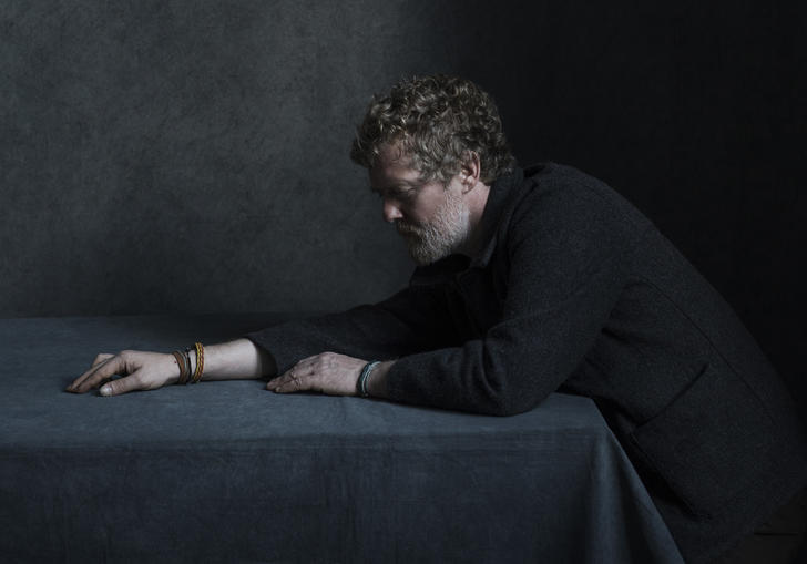 Glen Hansard stretching his right arm across a table. He is wearing leather bracelets.