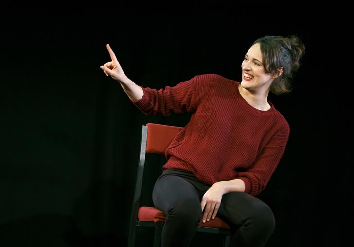Phoebe Waller-Bridge sitting on a chair in a dark space with her arm and finger pointing up