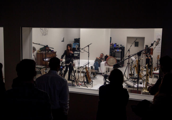 A group of musicians standing in a recording studio being watched by the producers in the next room