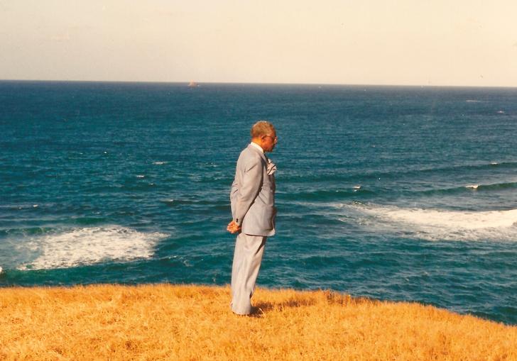 Aimé Césaire looks out to sea in a pale suit, with bright yellow grass beneath him 