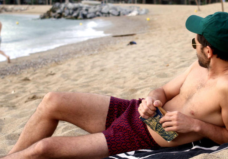 A man reclines on a beach in dark pink shorts and a teal cap, holding a book and looking at another man by the sea