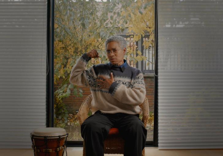 Glenn Copeland flexes a bicep in a grey and cream woolly jumper, in front of a window, next to a drum