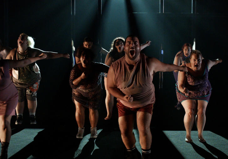 a dance troupe of dancers with larger bodies dances some choreography in a dark room 