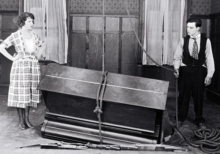 Buster Keaton and Sybil Seely stand in a room next to a piano which has crashed partially through the floor. 