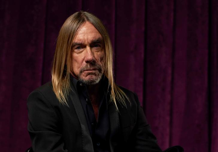 Iggy Pop, with long hair and a beard, is sitting on a chair in front of a velvet curtain