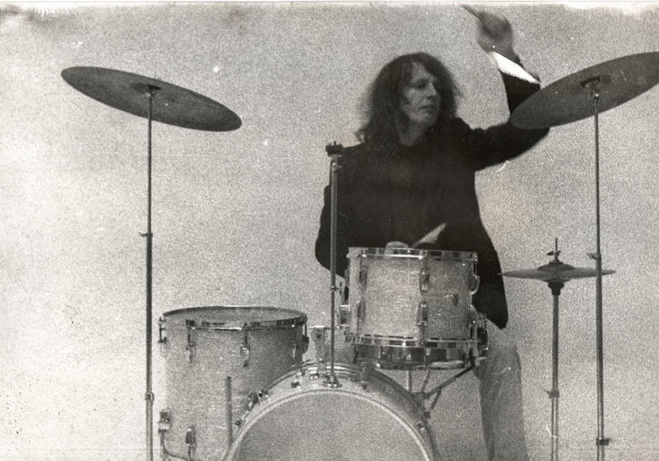 Klaus Dinger playing a classic drum kit against a bare wall