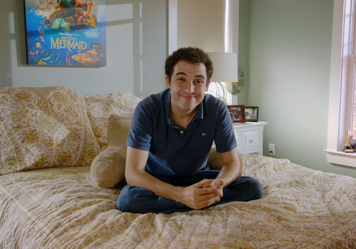 A young man sits on a bed smiling, with a poster of the Little Mermaid behind him
