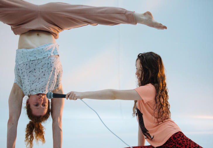 adult is in a handstand with her legs in splits, with a girl on the floor holding a microphone to her mouth