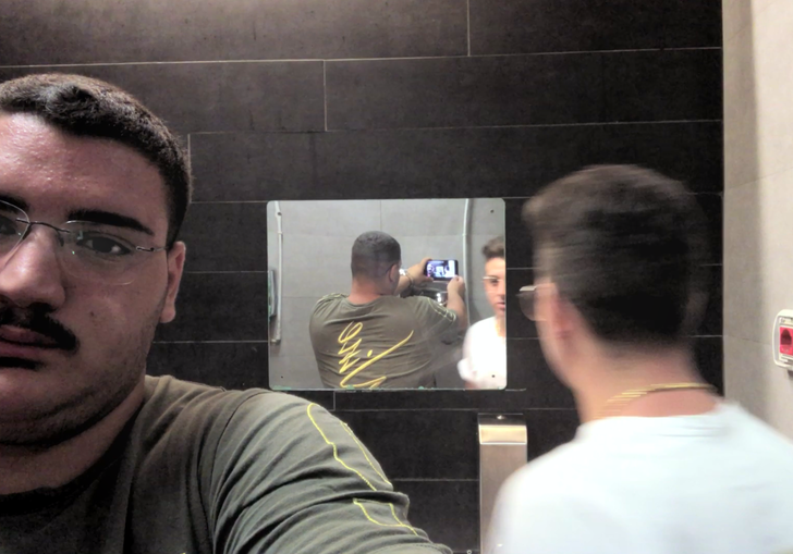A man takes a selfie in a bathroom and we see this reflected in a mirror opposite, while a friend looks directly into the mirror