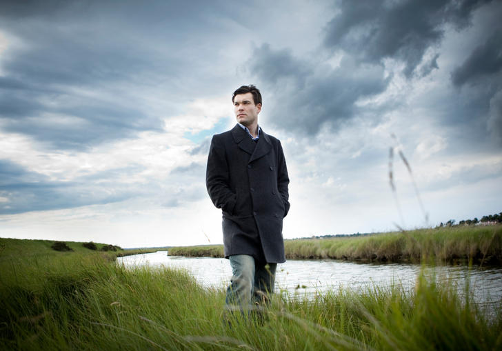 An image of Ryan standing on the grassy river bank, a dramatic grey sky overhead 
