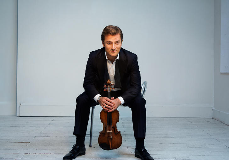 Renaud sitting on a chair in a white room with a cheeky grin on his face, clutching his violin