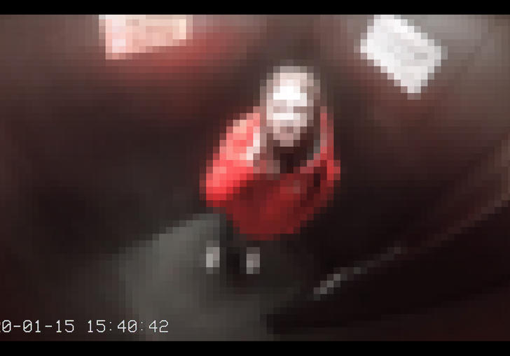 A pixelated view from a CCTV camera of someone looking up at the camera in a small room. They are wearing a red coat and they have brown hair.