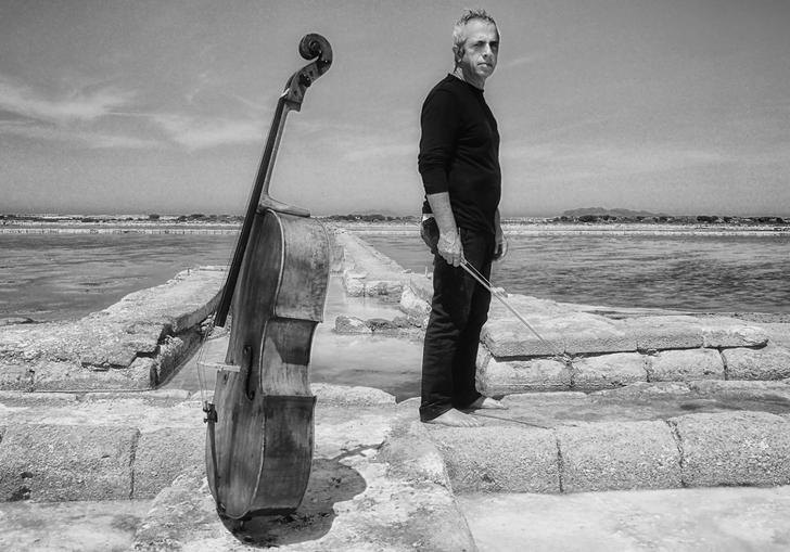 A black and white image of a pensive Giovanni standing barefoot on some ruins in a dry and barren wasteland, his cello standing upright close by