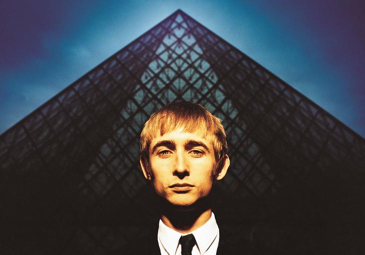 Neil Hannon in a black and white suit and tie standing in front of a blue pyramid
