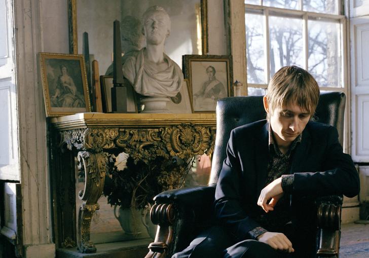 A colour photo of Neil Hannon lounging in a chair in a slightly dilapidated stately home, with antique furniture in the background