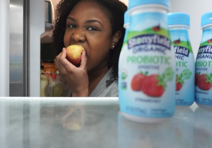 a woman eating an apple looks into her fridge and sees some probiotic yogurts
