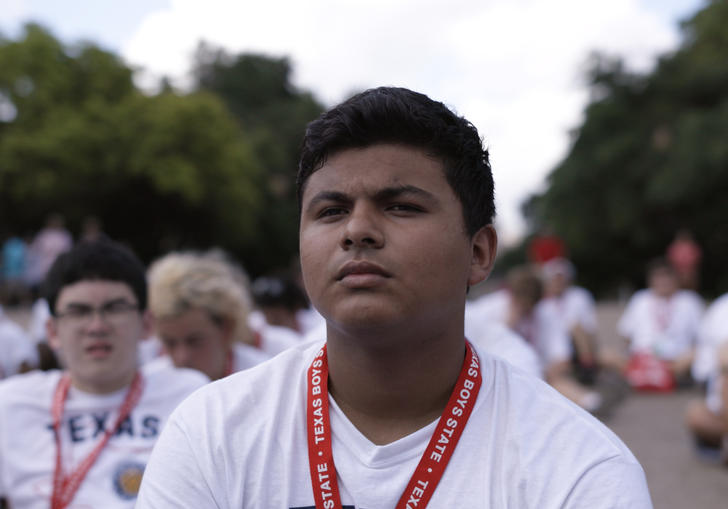 a boy in a white t shirt sits with many others behind him