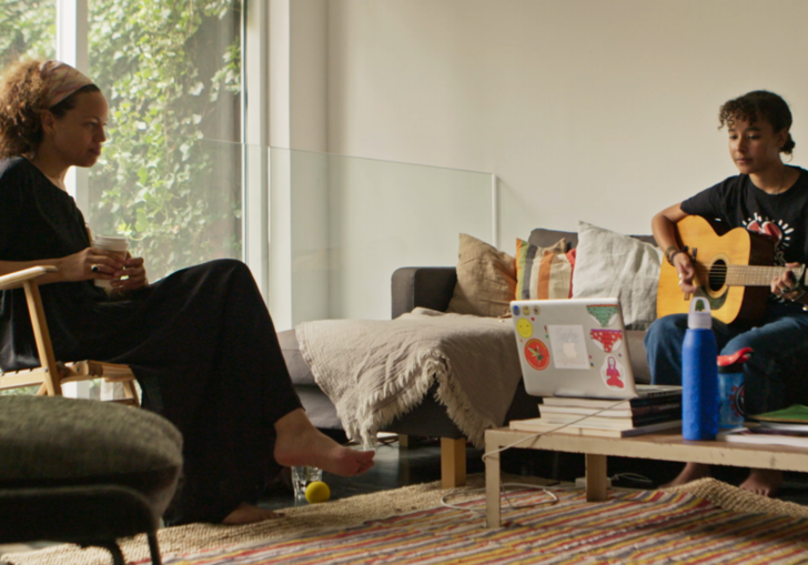 two women sit across from each other in a room, one holding a guitar