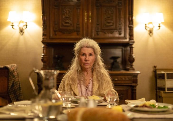 an old woman sits at the head of a long table with her long white hair cascading down