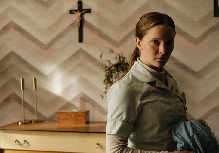 A woman stands looking toward the camera in front of a cross