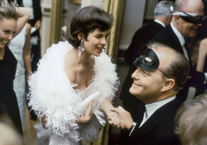 Capote sitting in a room with a mask on his head surrounded by people