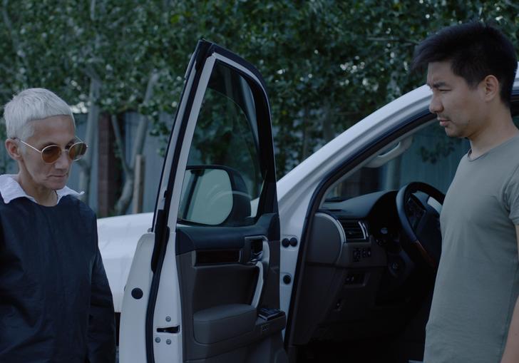 a man and a woman stand in front of a car looking at each other