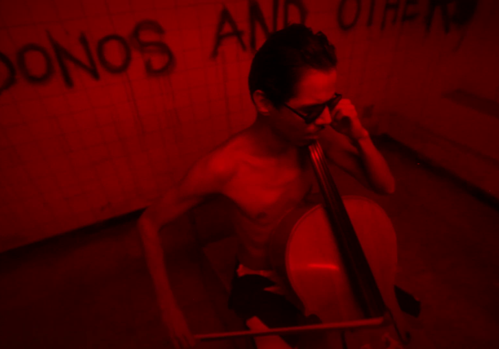 a man plays a cello in a dark room full of red light and graffiti on the wall