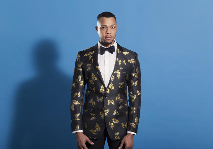 Davone Tines in a suit, in front of a blue wall