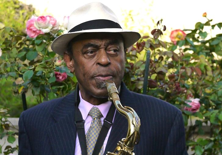 Archie Shepp playing a saxophone. He is wearing a black suit with a white hat and standing in front of a rose bush.