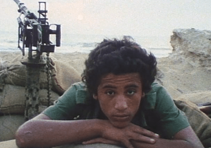 A young man lies on sand bags with a automatic gun behind him