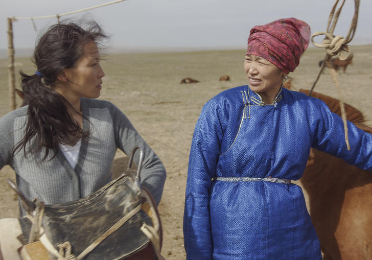 Two sisters in the desert look at one another, standing next to livestock