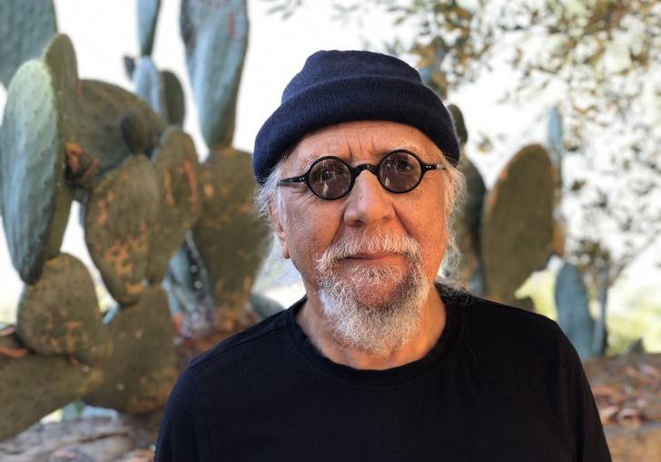 Charles Lloyd in a black beanie hat and small round sunglasses, standing in front of a large cactus