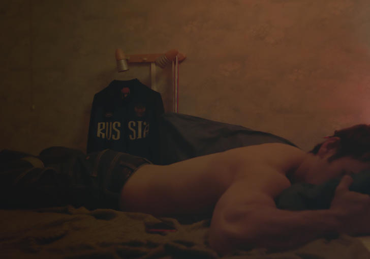 A man lies in on a bed belly down, in a dimly lit room, with a Russia hoodie in the background