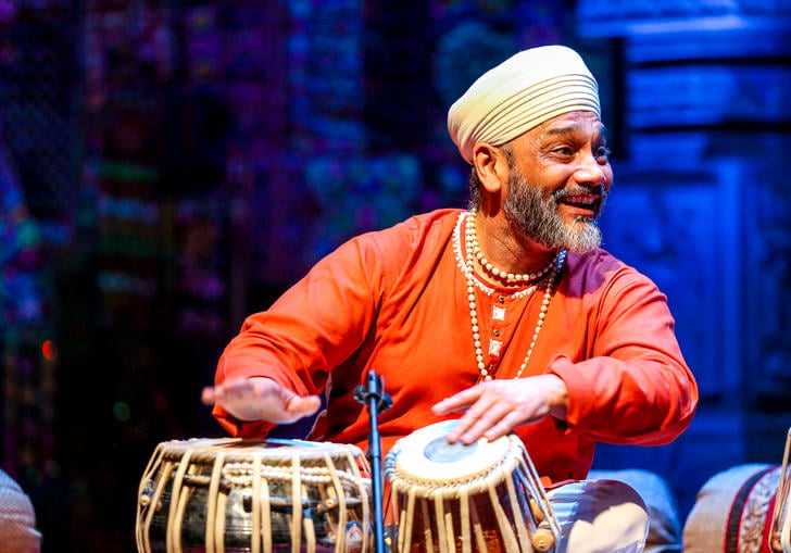 Sukhwinder Singh playing the tabla and smiling to his left. He is wearing an orange top, beaded necklaces and a white turban.