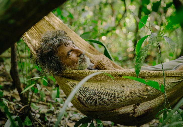 A man with grey hair and a beard lies in a hammock in foliage, in a still from El Father Plays Himself