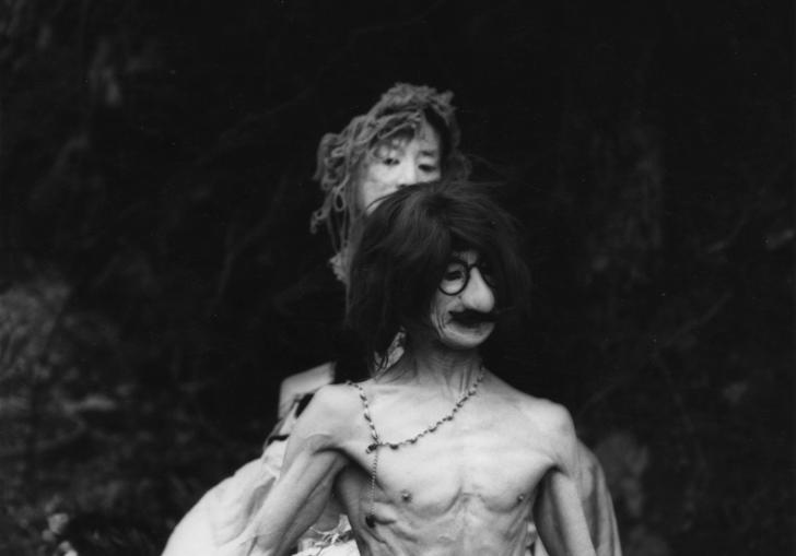 a man wearing an ugly mask stands topless with a woman behind him