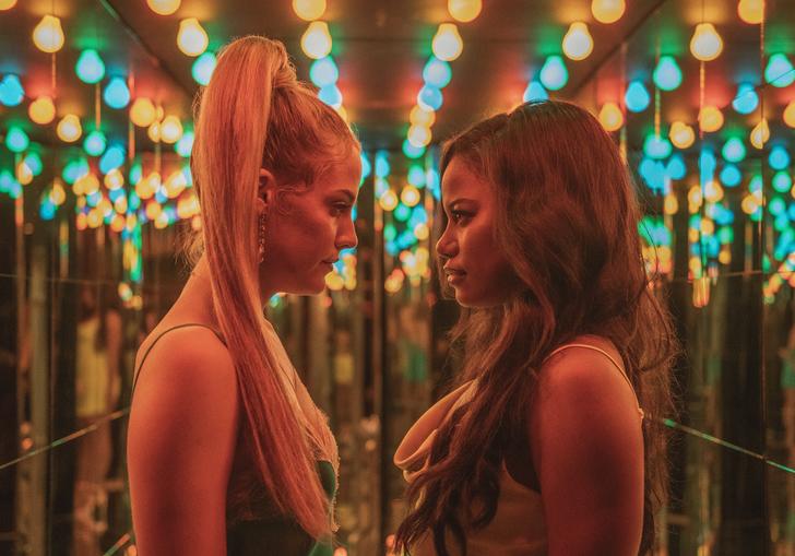 Riley Keough and Taylour Paige look directly at each other in a hallway of mirrors with multicoloured lights above