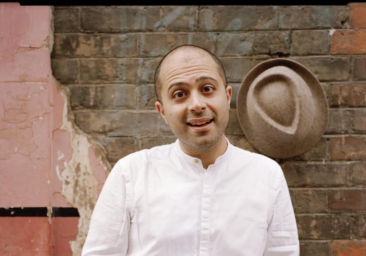 Mahan Esfahani smiling in front of a wall, with his hat hanging on the wall behind him