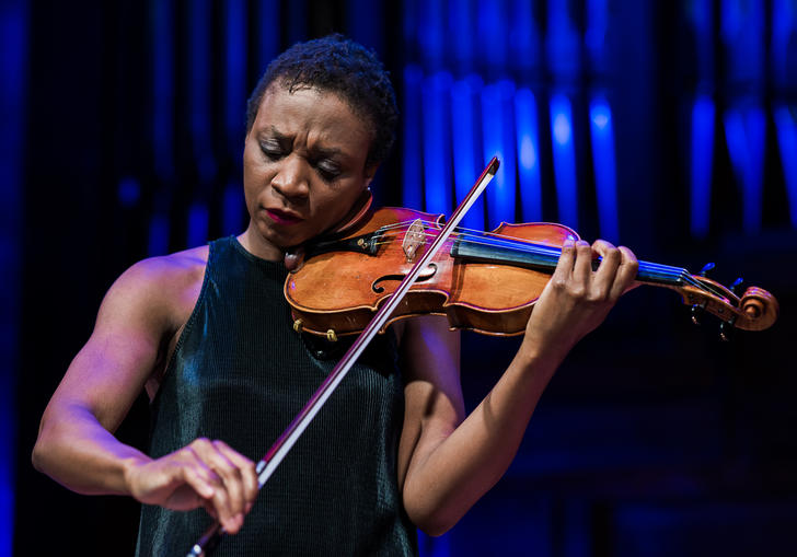 Tai Murray stands playing the violin, in front of organ pipes with a blue light on them