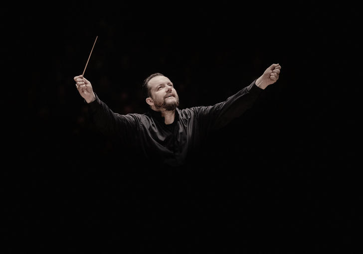 Andris Nelsons with his arms raised. He's holding a conductor's baton in his right arm, and he appears to be coming out of the black background. 