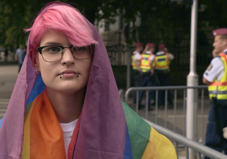 Tobi, a young person with pink hair, is draped in the rainbow flag in front of a few police officers in a still from Colors of Tobi