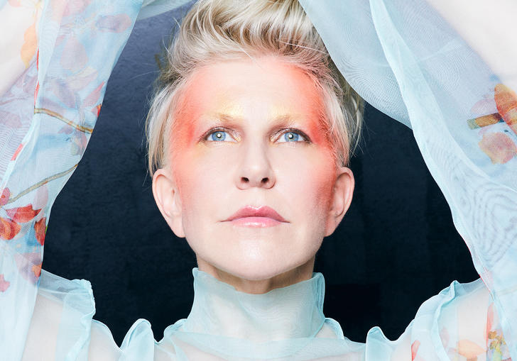 Joyce DiDonato in a blue floaty top with her arms above her head, with red face paint on her temple and the sides of her face