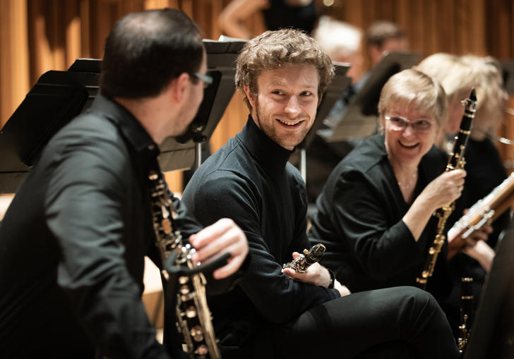 Two members of Britten Sinfonia smile at another member, who has his back to the camera. They are all on the Barbican stage, holding their instruments.